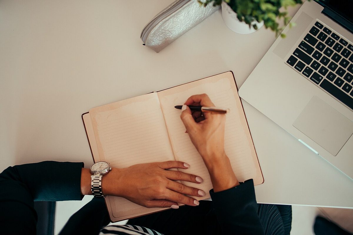 A personal-finance blogger writing notes in a notebook beside a laptop at a desk