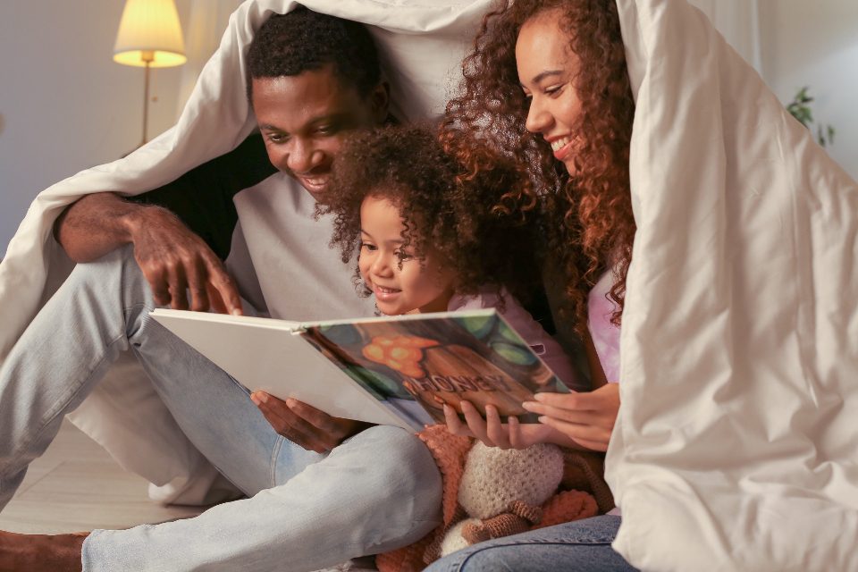 Two parents and a young child reading a picture book together under a blanket